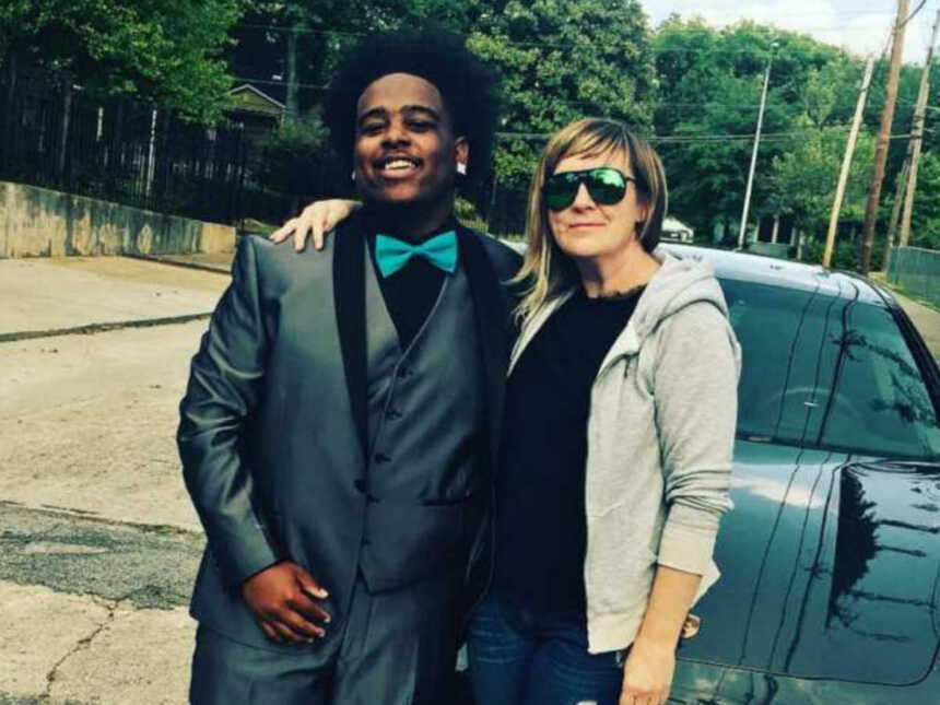 Woman holding teen in gray prom suit in front of porsche