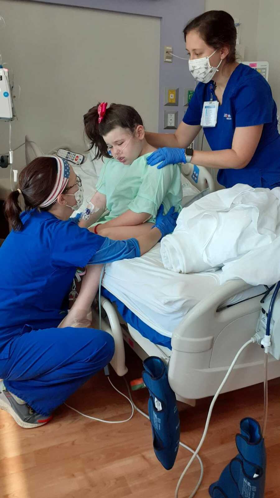 Two nurses holding up girl in hospital bed