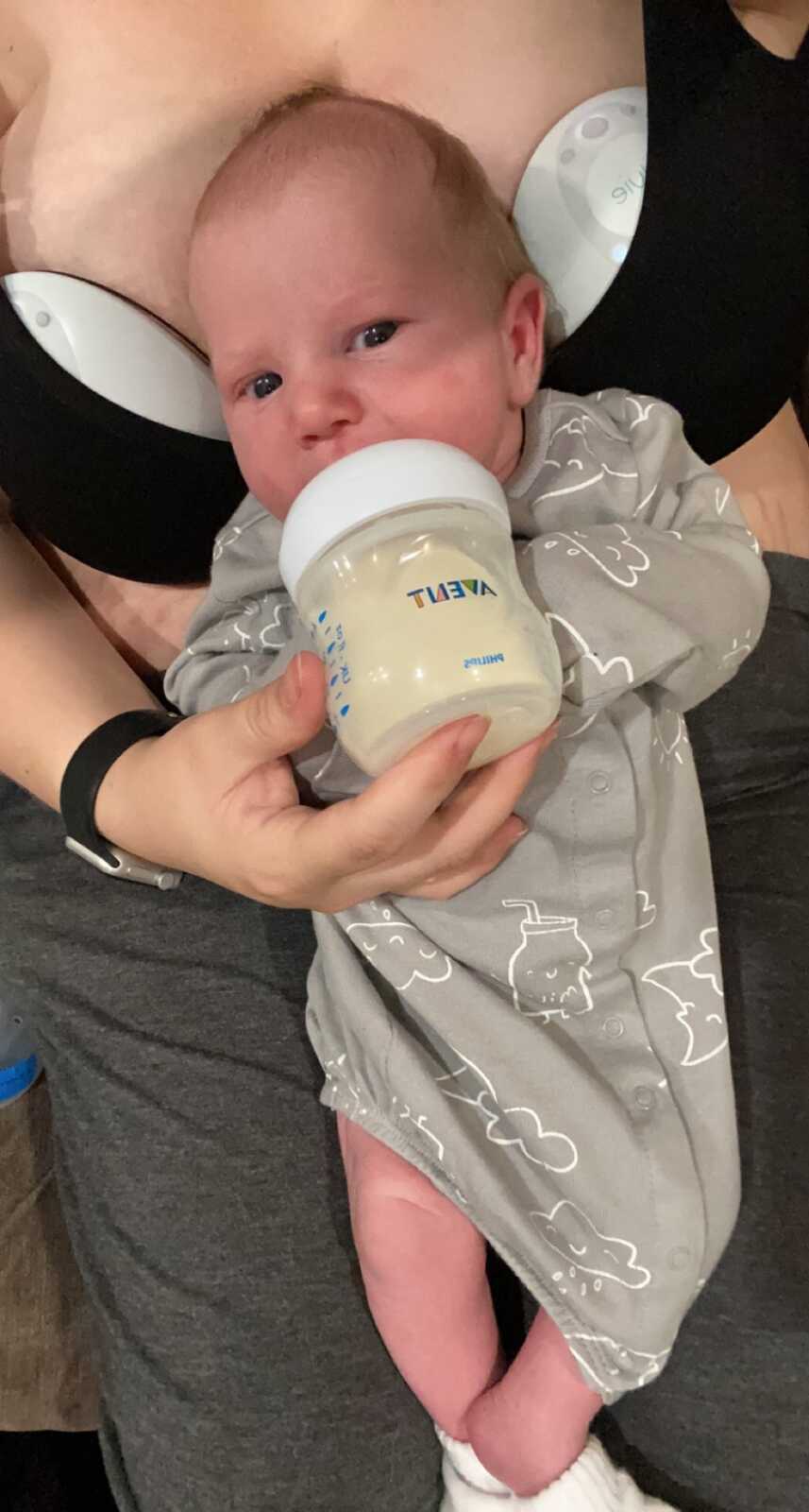Infant in gray jumper sips bottle in mother;s lap