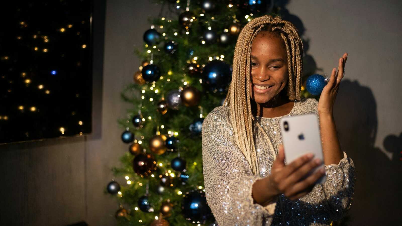 a woman chats on a facetime call on her phone in front of a decorated Christmas tree