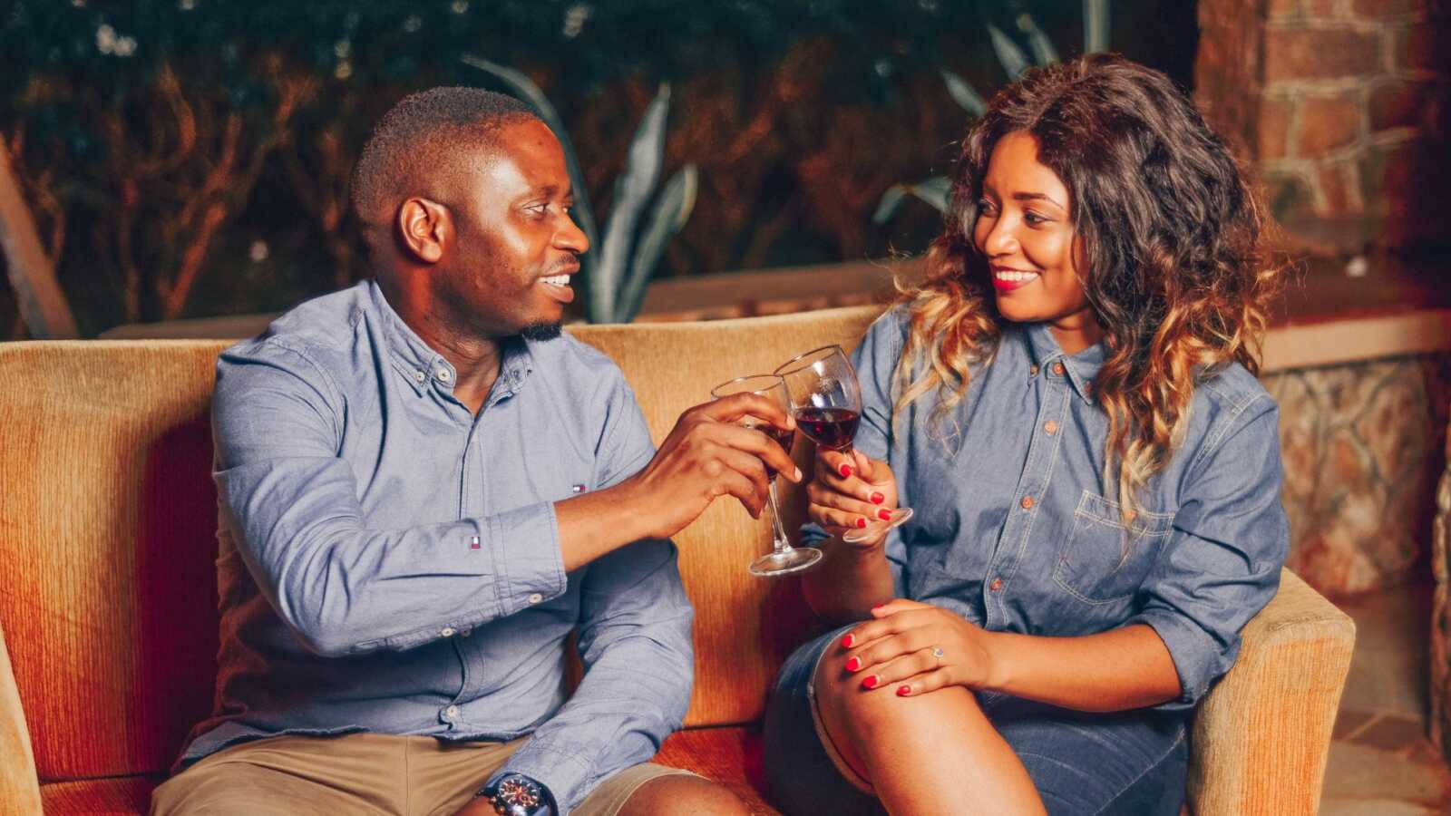 a man and woman sit together on a couch and cheers with glasses of red wine