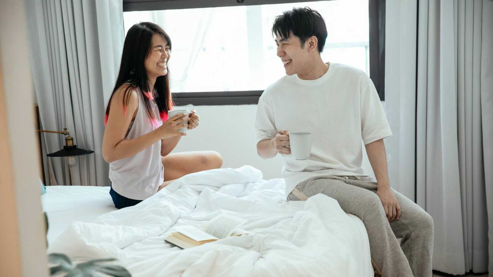 a couple sits together on an unmade bed laughing while holding coffee cups