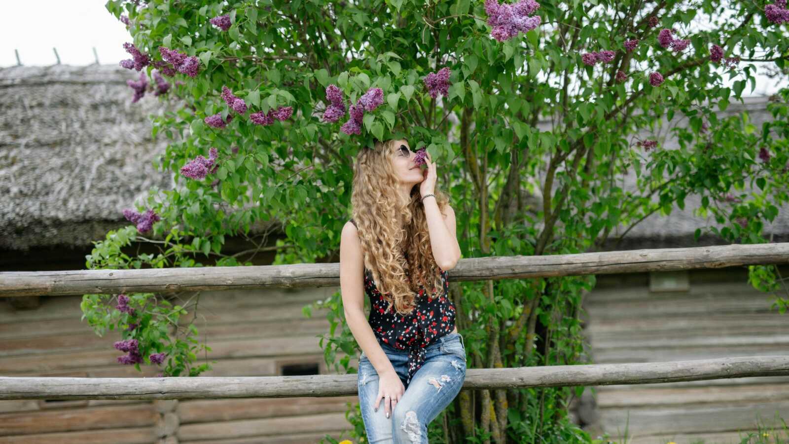 a woman sitting on a fence smelling a purple flower on a tree behind her