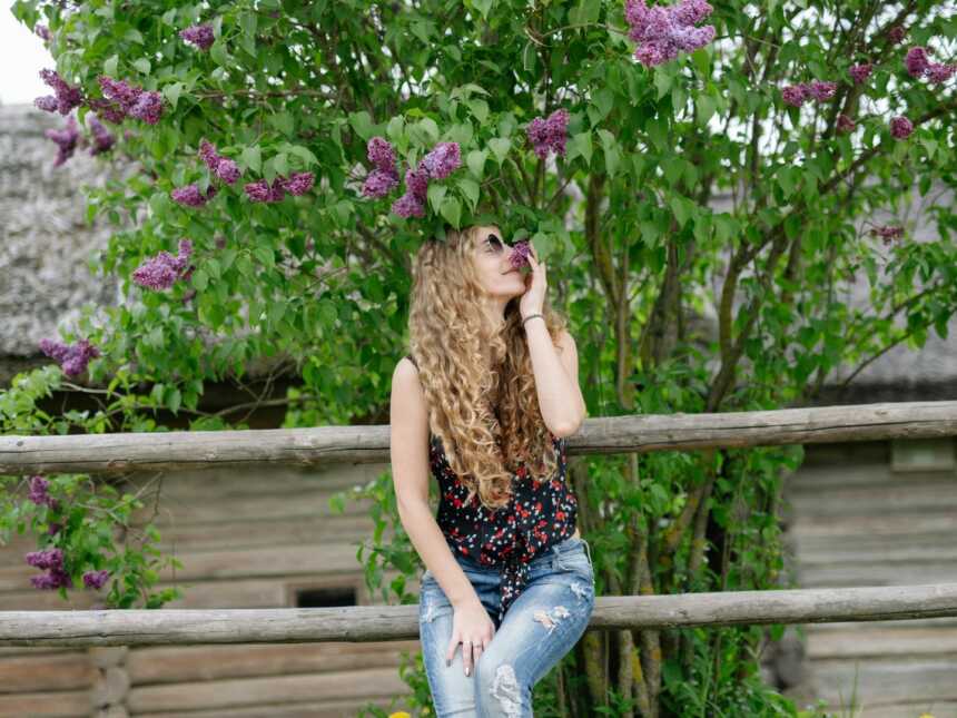a woman sitting on a fence smelling a purple flower on a tree behind her