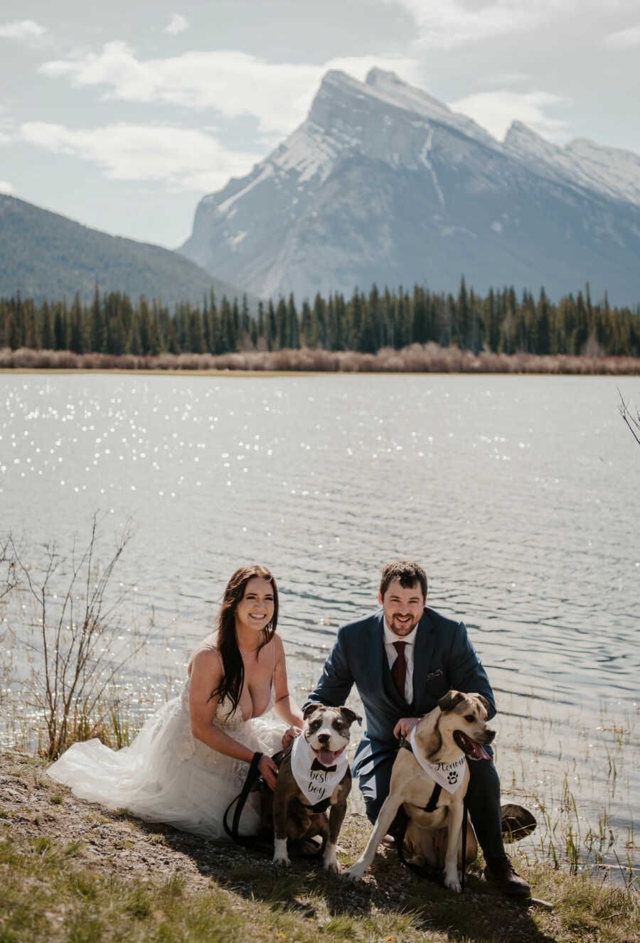 Bride and groom smiling with two dogs beside lake